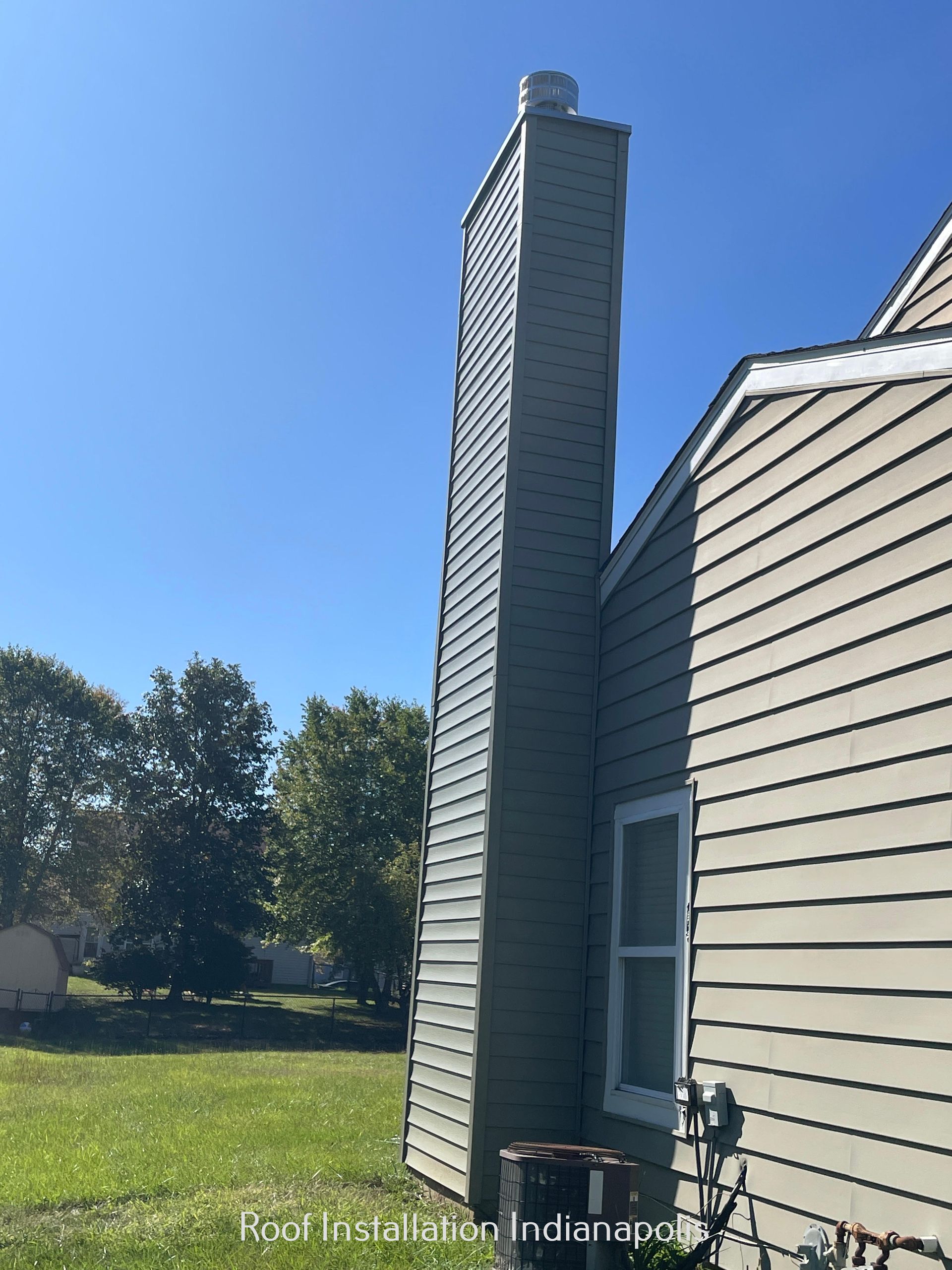 A tall, tan-sided chimney stands next to the exterior wall of a house under a clear blue sky.