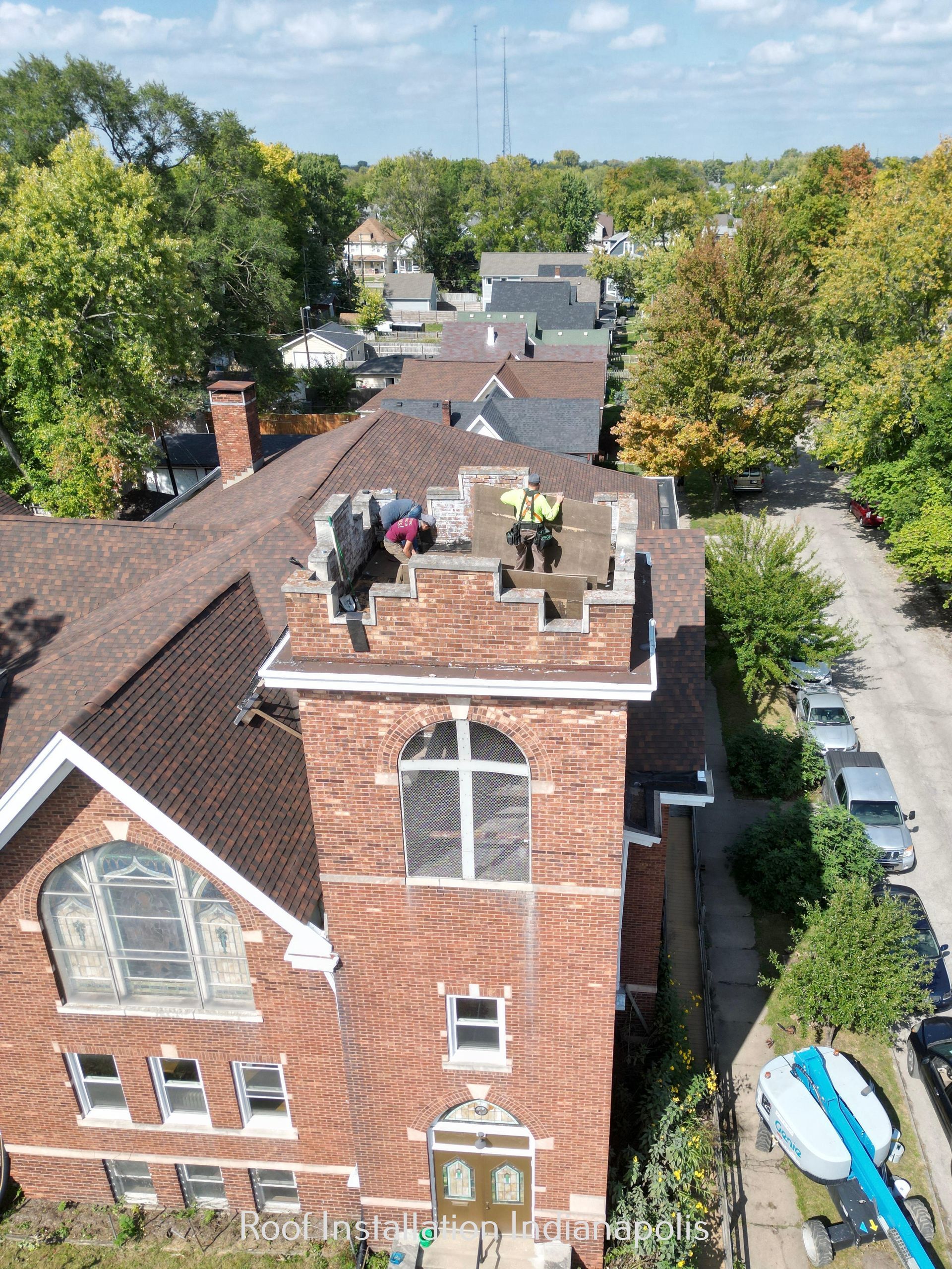 Two workers perform construction on the brick tower of a church roof, with a street and trees visible below.