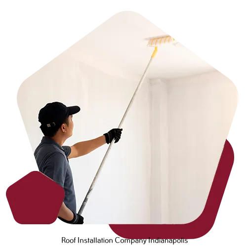 A person wearing a hat and gloves uses a long-handled roller to paint a ceiling white.