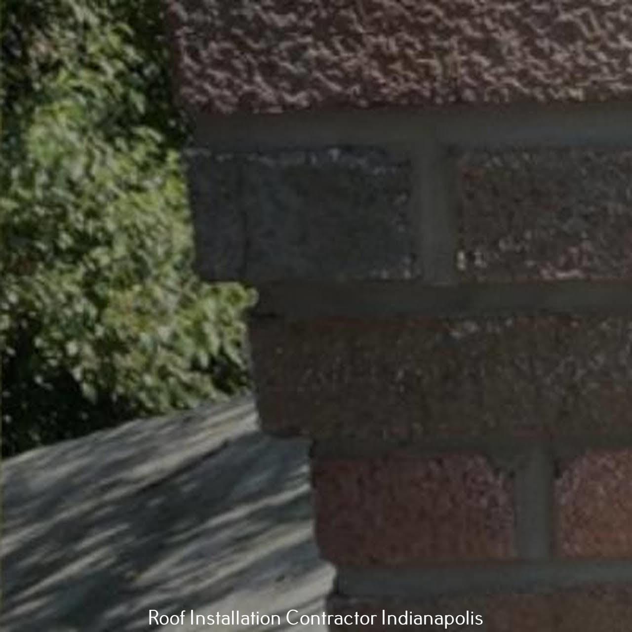 A close-up of a textured red brick chimney corner meeting a shingled roof with foliage in the background.