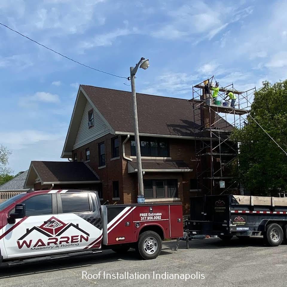 A Warren construction truck and trailer parked in front of a brick house with workers on scaffolding repairing the roof.