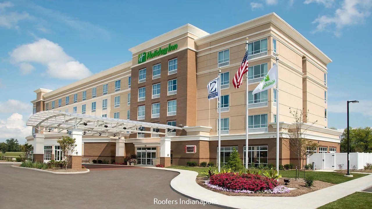 A beige Holiday Inn hotel with a modern entrance canopy, a flag display, and landscaped flowers on a sunny day.