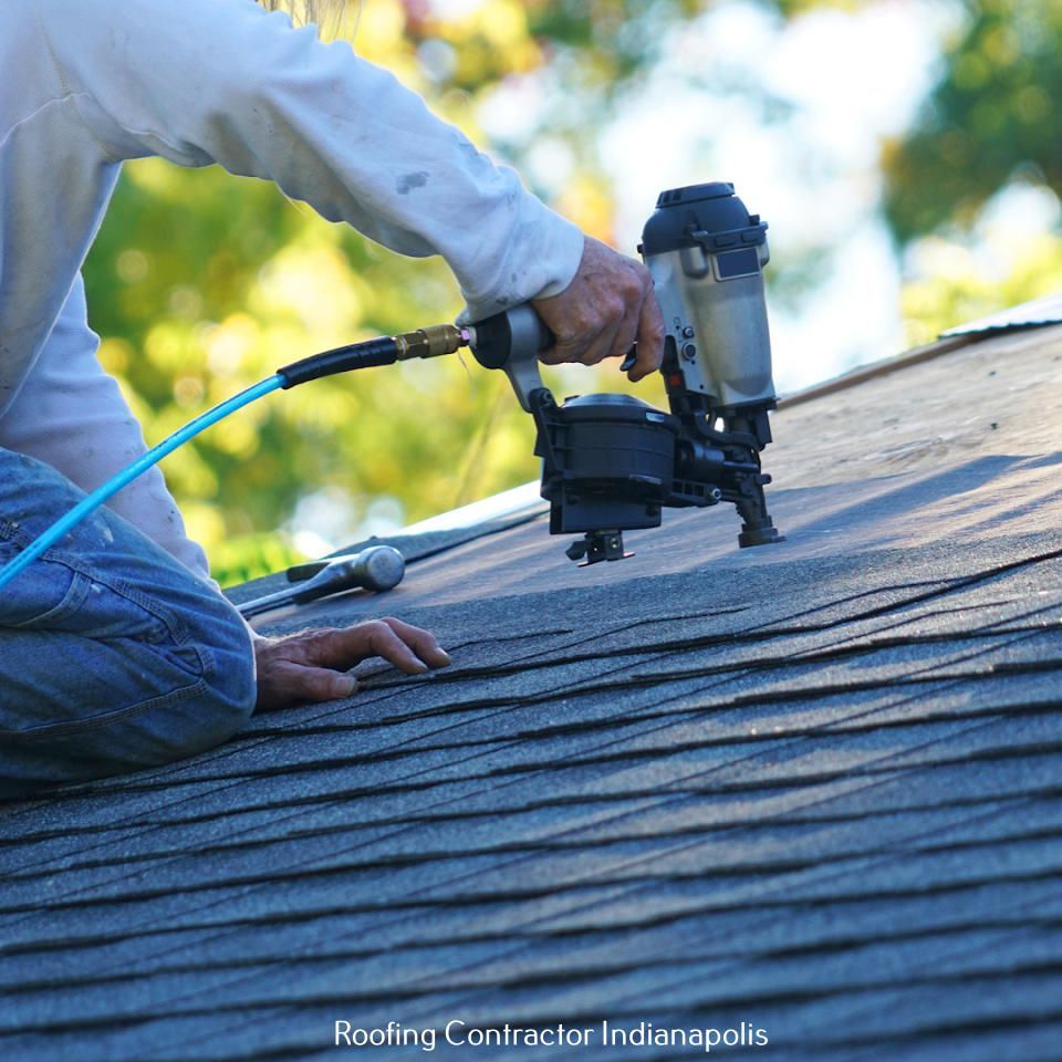 A person uses a pneumatic nail gun to install dark asphalt shingles on a residential roof.
