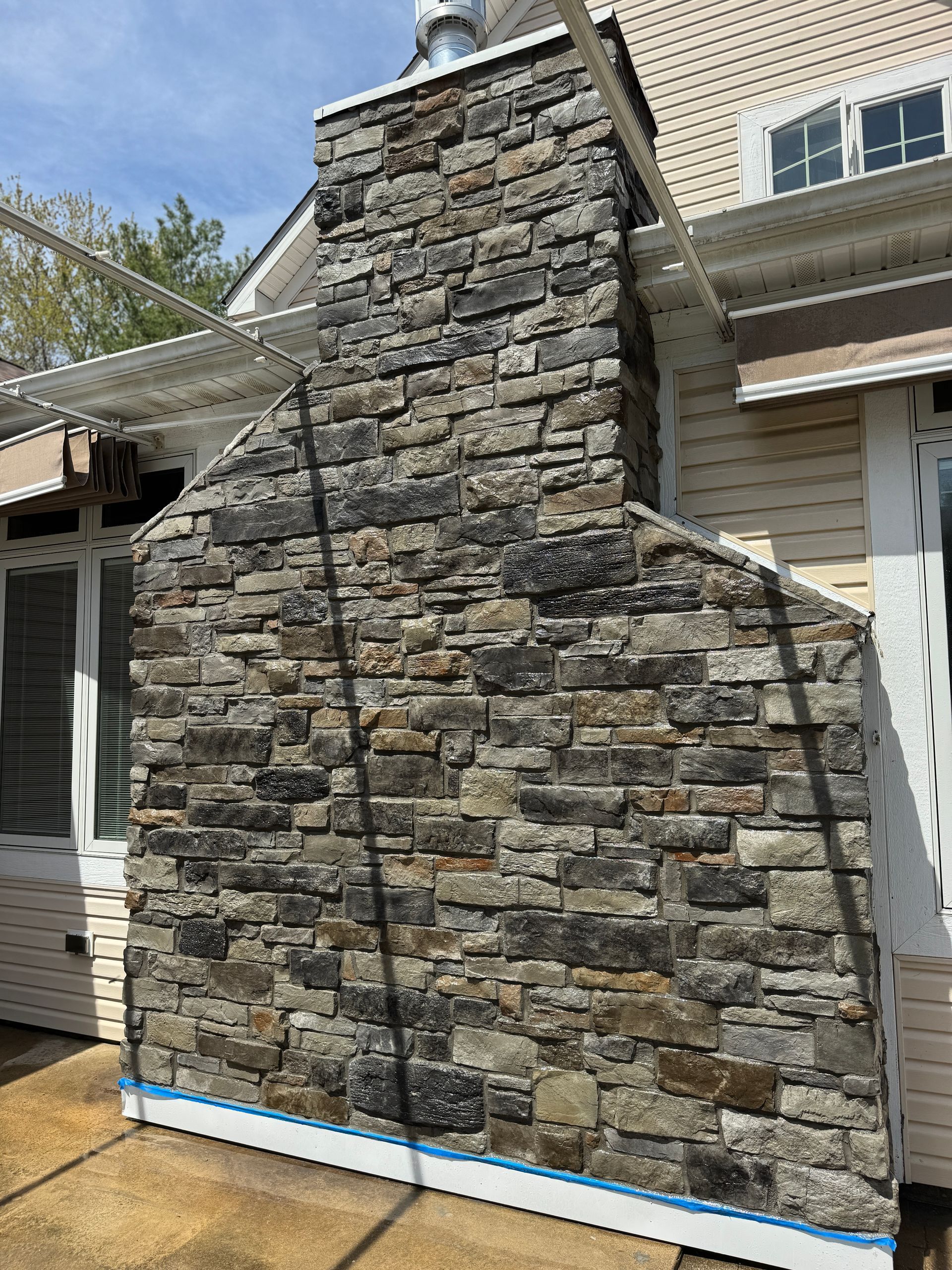An outdoor stone fireplace chimney attached to a light-colored, siding-covered house on a sunny day.