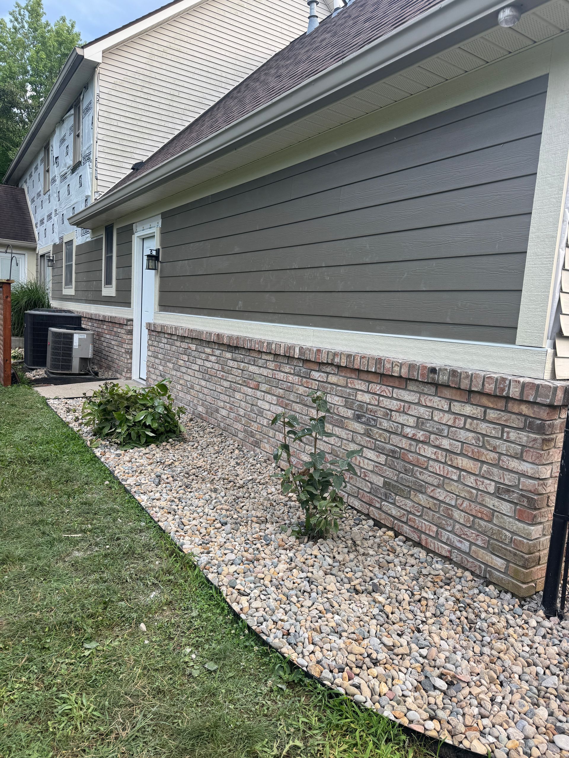 A side view of a house exterior featuring dark horizontal siding above a brick foundation, bordering a stone landscape bed.