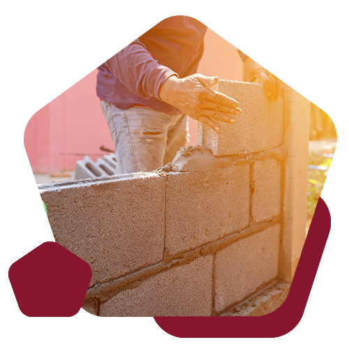 A worker in jeans and a long-sleeved shirt carefully laying concrete blocks to build a wall on a sunny day.