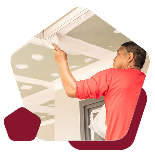 A person in a red shirt applies white filler to a drywall ceiling seam in a residential room.
