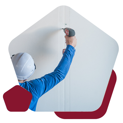 A worker in a blue shirt and white hard hat uses a power drill to fasten a white panel to a wall.