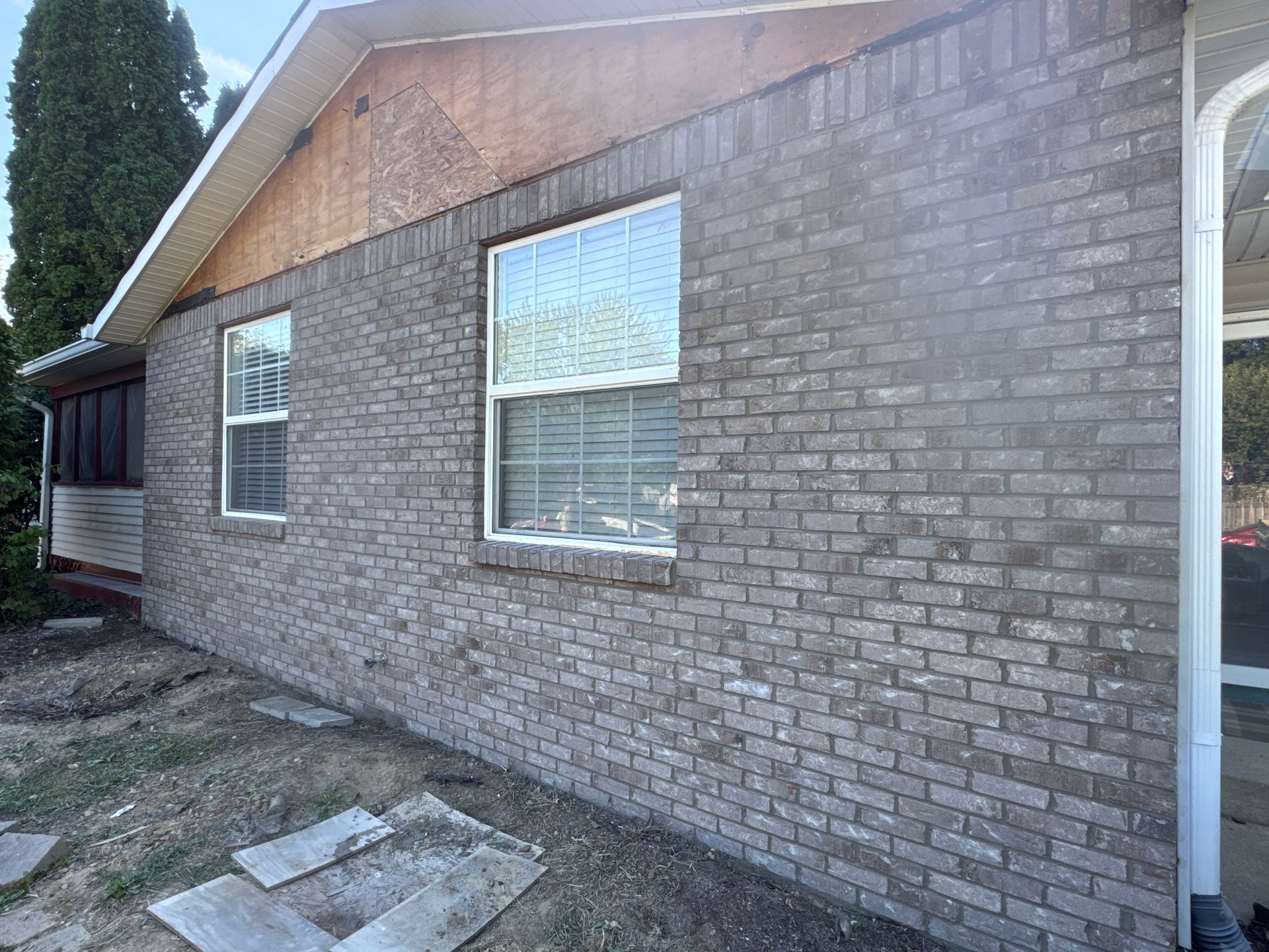 Side view of a house exterior featuring a brick wall, two windows, and an unfinished wooden triangular gable section.