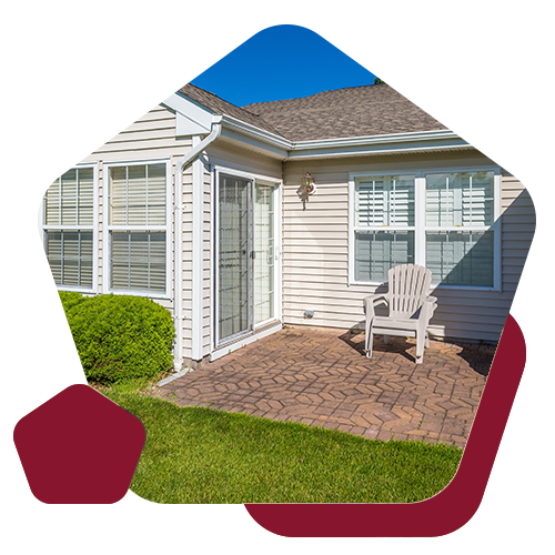 A tan vinyl-sided house with a stone patio, sliding glass door, and white lawn chair on a sunny day.