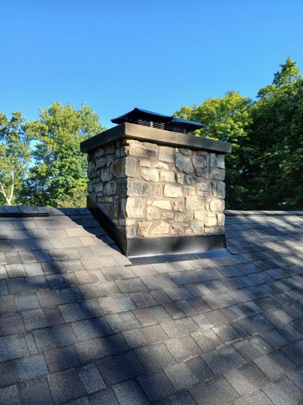 A stone chimney sits on an asphalt shingle roof against a clear blue sky and green trees.