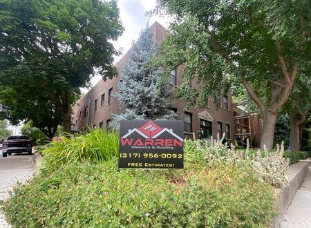 A Warren Property & Roofing sign sits in front of a brick building surrounded by green trees and landscaping.