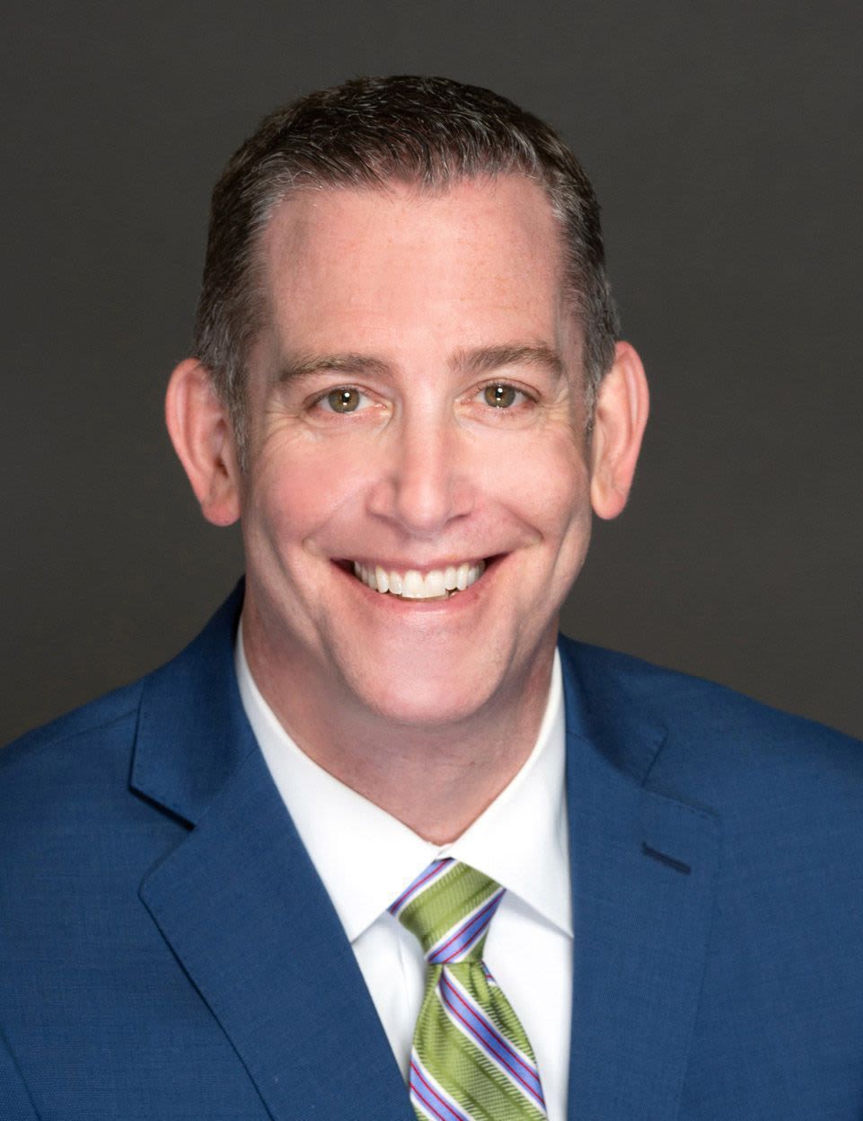 Man in blue suit and green tie smiles, with a plain gray background.