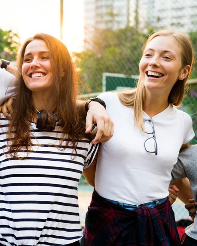 Two smiling friends standing arm-in-arm outdoors, one in a striped shirt and one in a white shirt and plaid skirt