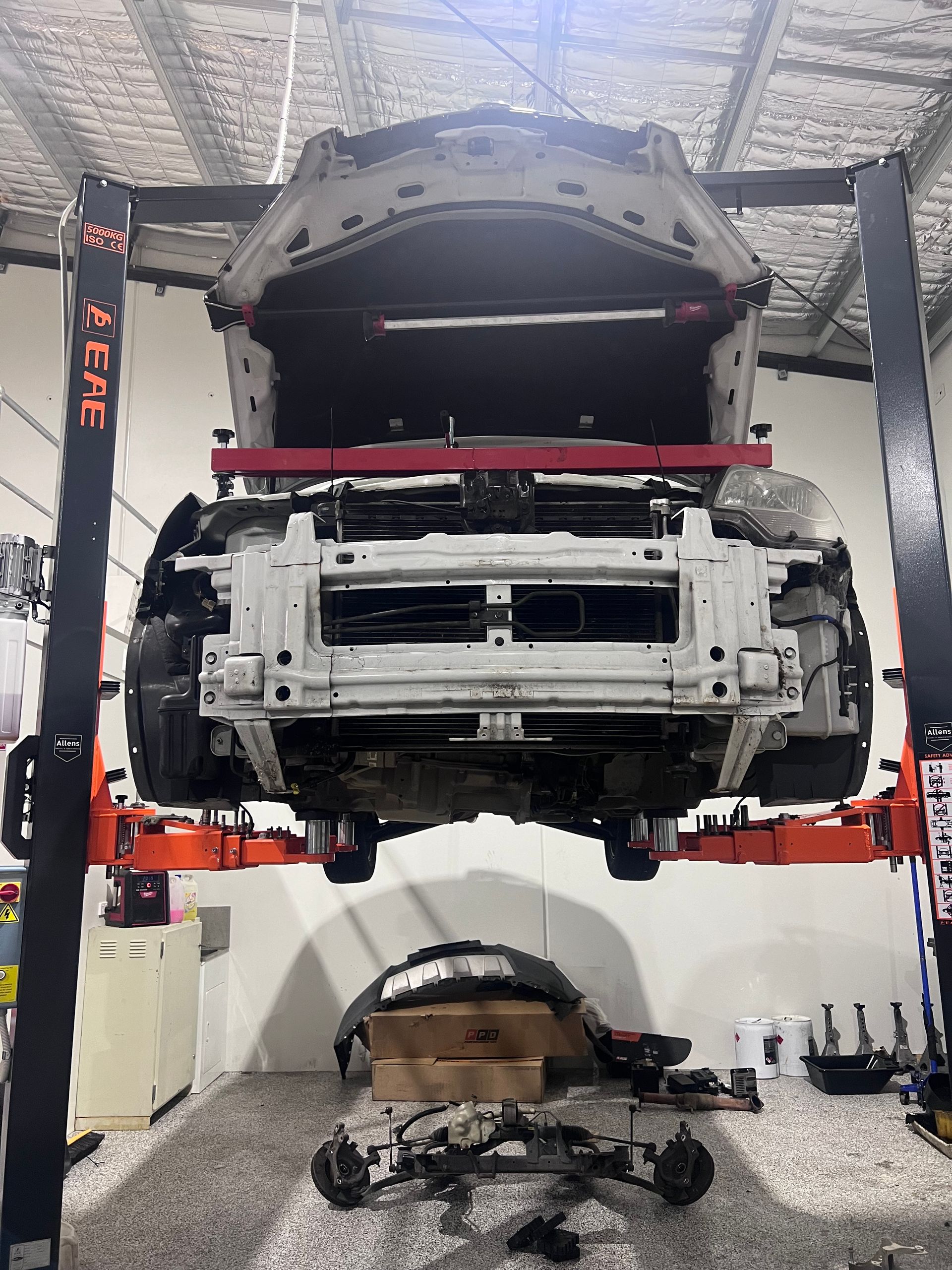 A car hoisted on a lift in a garage, undergoing repair. White car parts visible; tools on the floor.