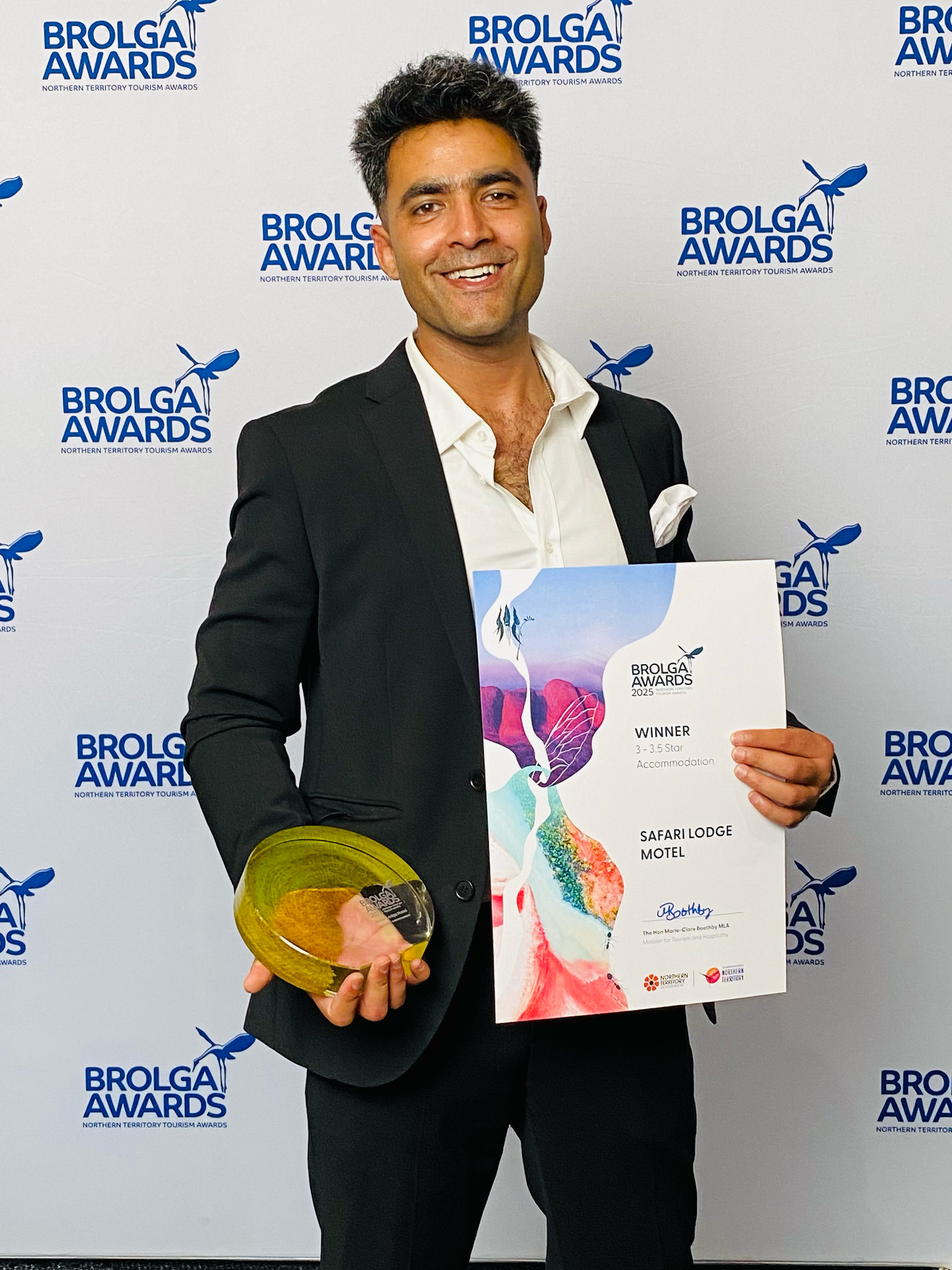 Man in dark suit holding an award plaque and trophy at the Brolga Awards backdrop