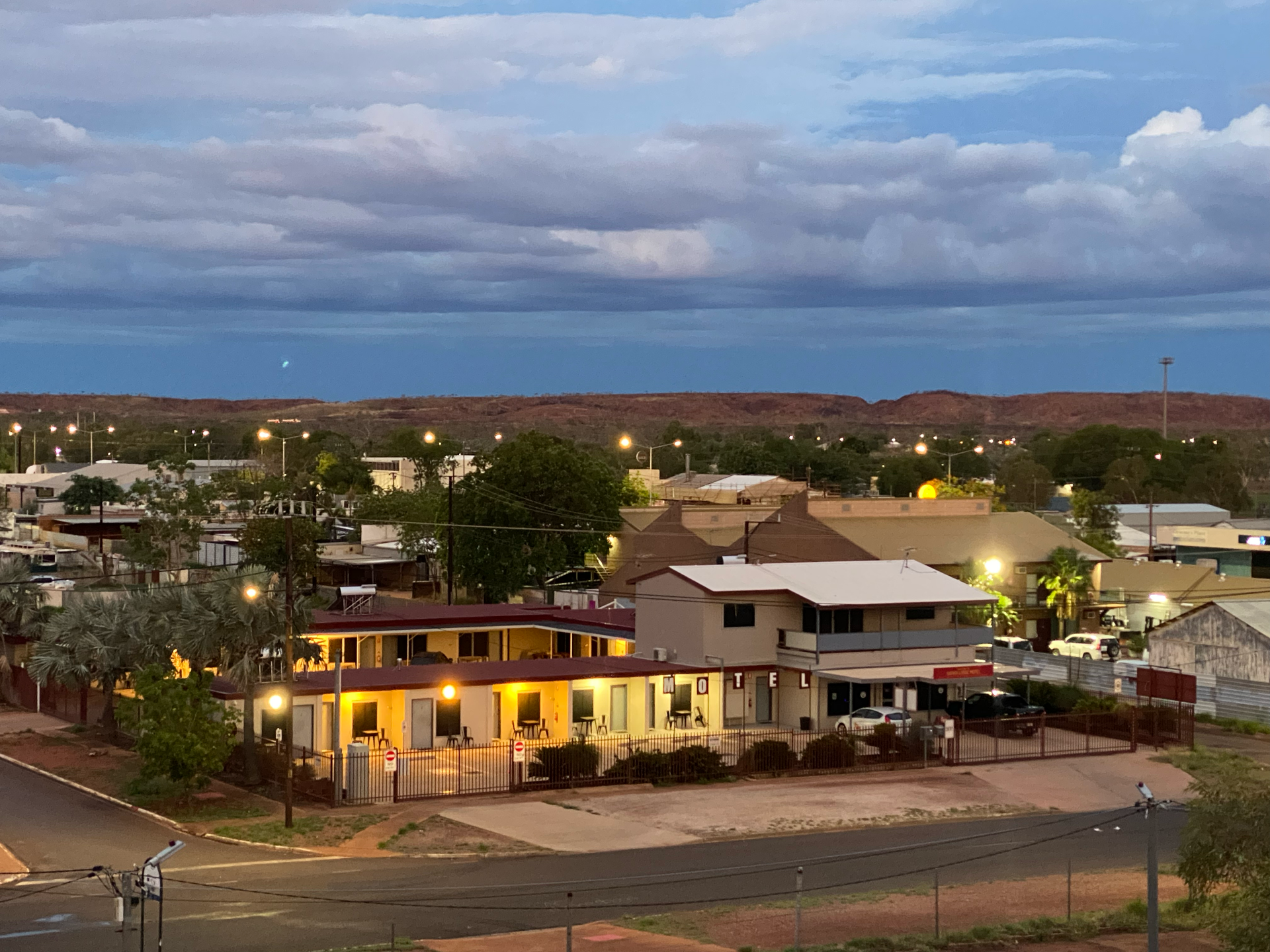 Sunset over a small town with lit buildings, trees, and distant hills under a cloudy sky