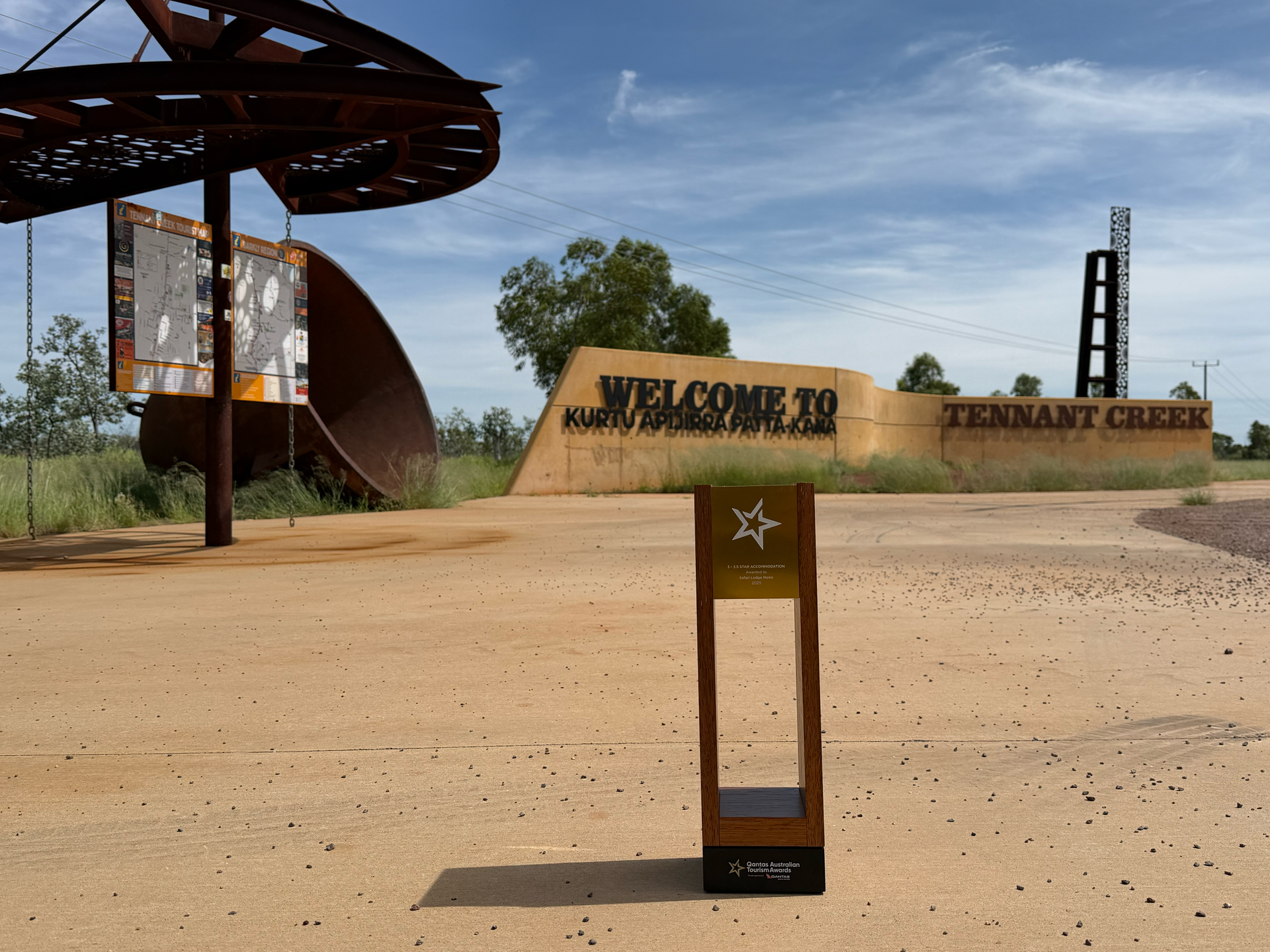 Sculptural entrance sign in a desert landscape under a blue sky, with a metal canopy and sandy ground