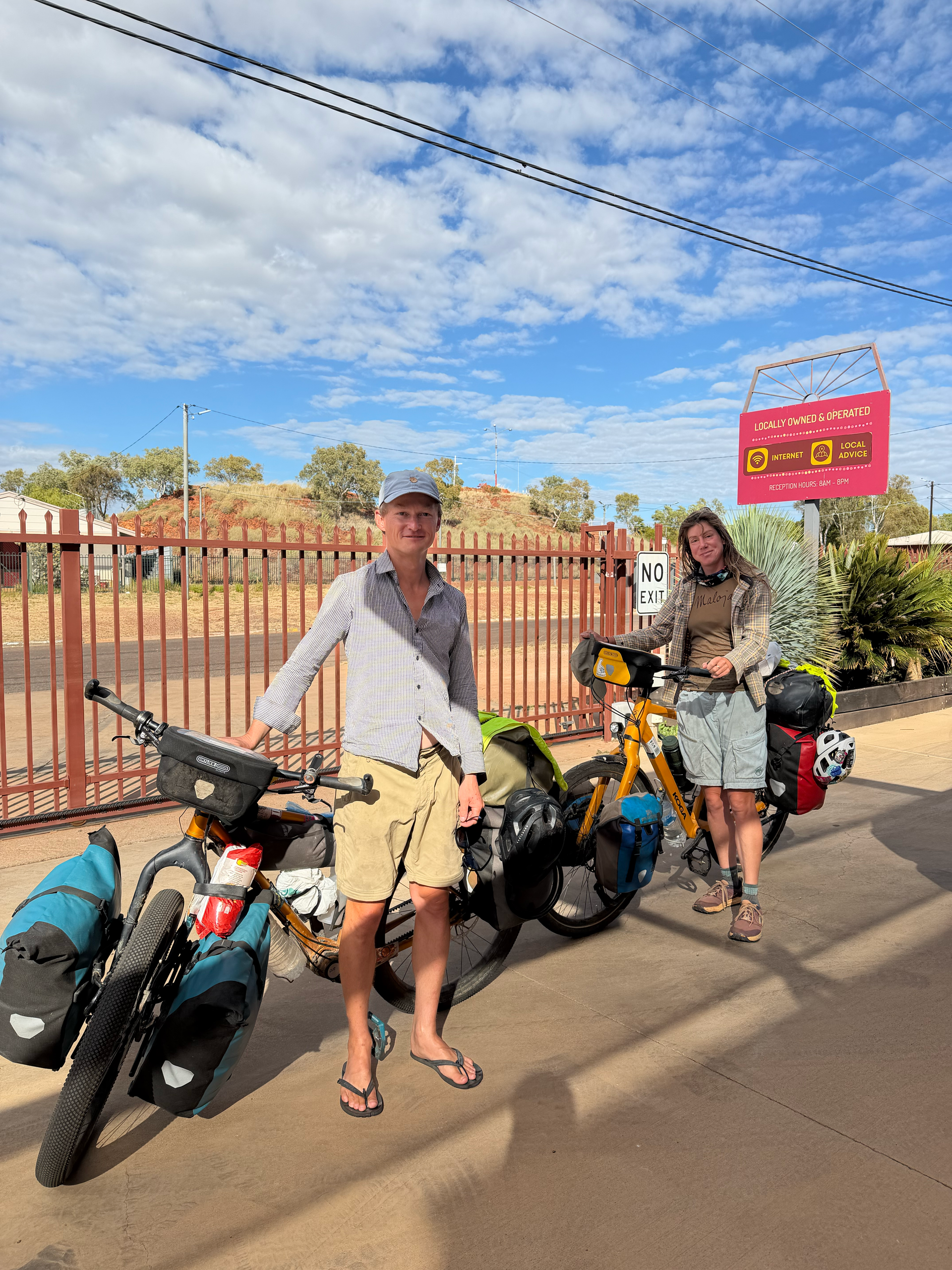 Two cyclists standing with loaded touring bikes on a sunny road beside a fence and sign