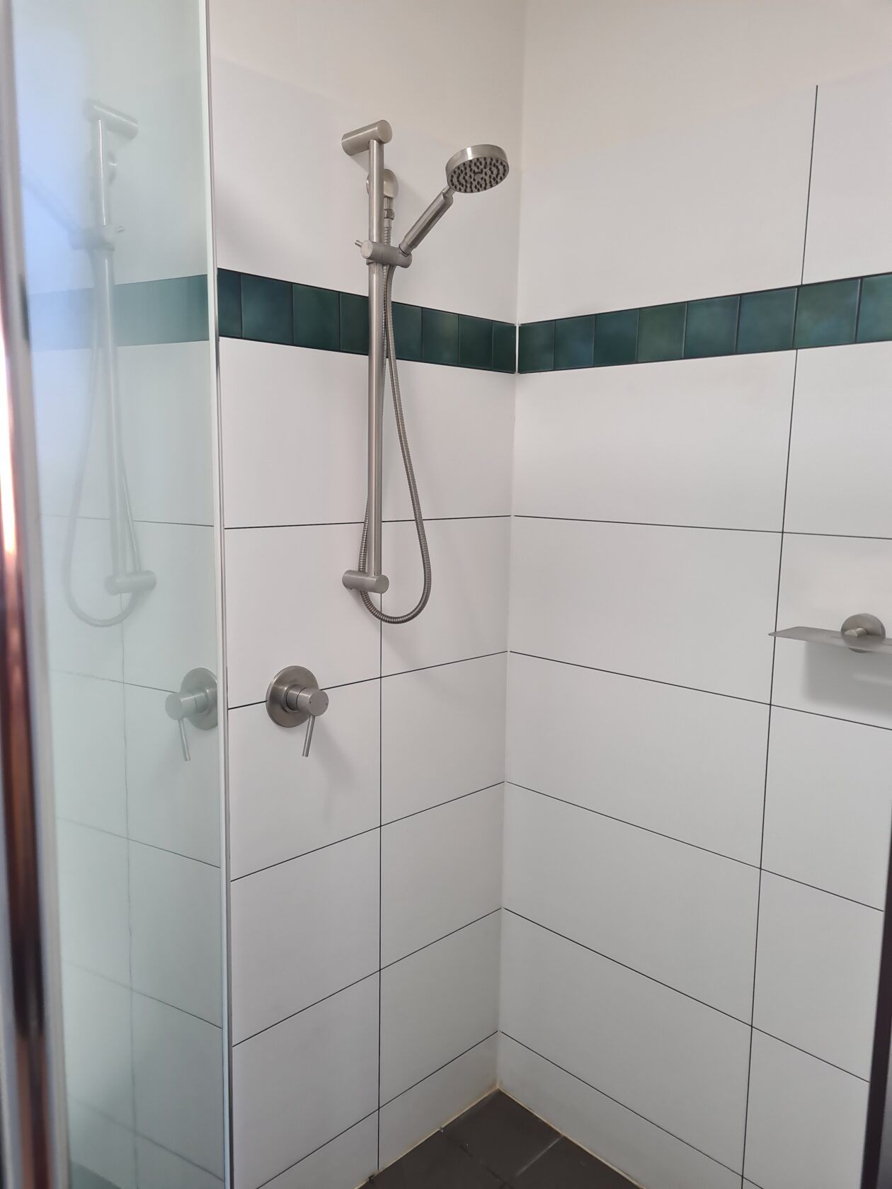 A shower with white tiled walls, a glass door, and a silver shower head and faucet. A dark green stripe runs along the top of the tile.