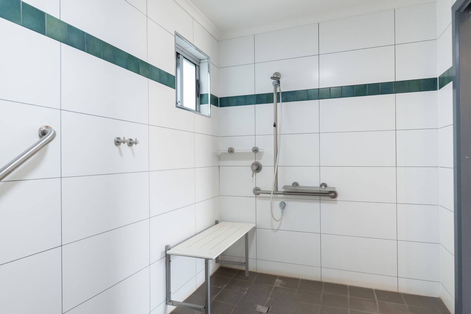 Accessible shower with white tiled walls, a bench, grab bars, and a hand-held shower head.