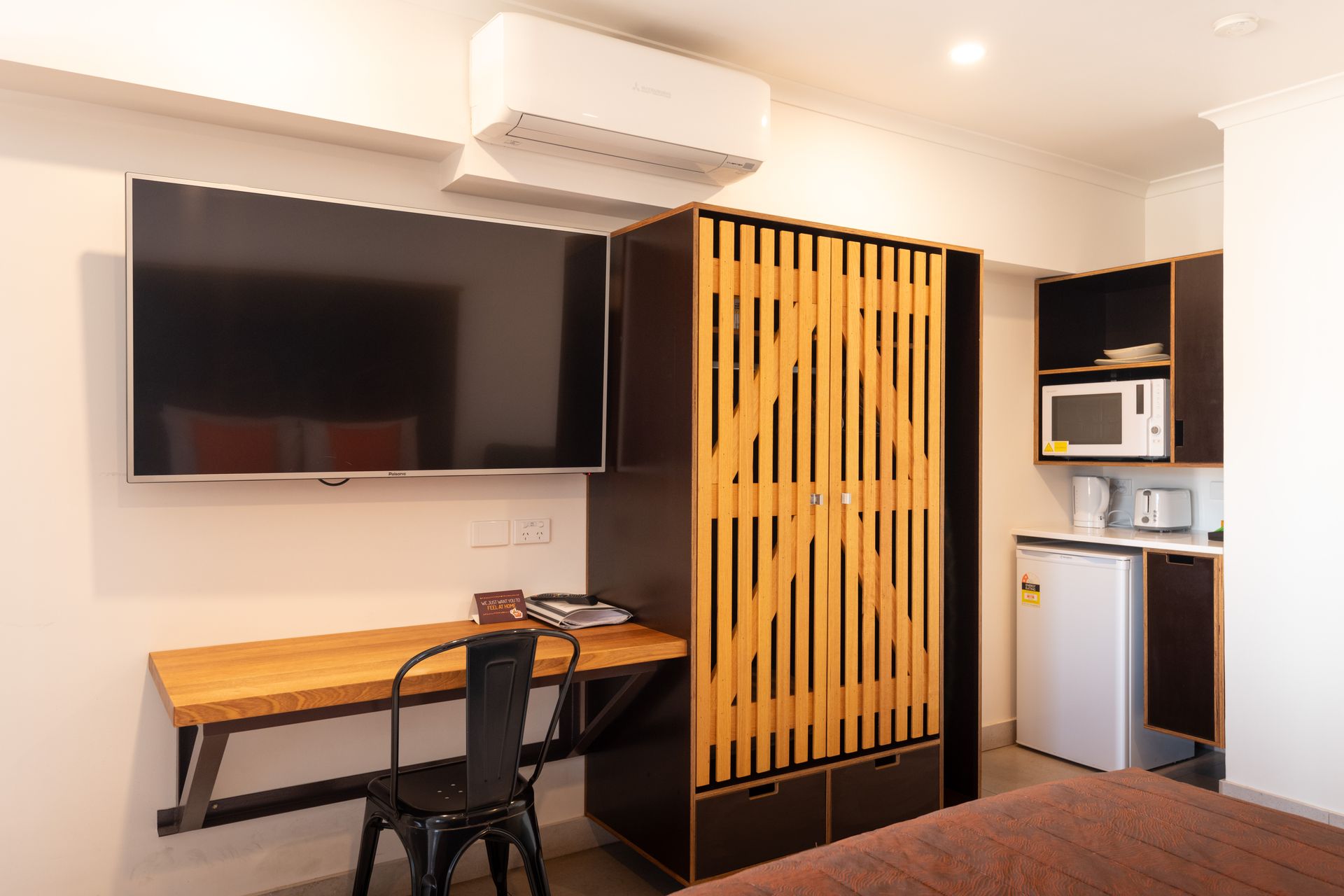 Small hotel room with a mounted TV, desk, and wardrobe, with a microwave and mini-fridge in the corner.