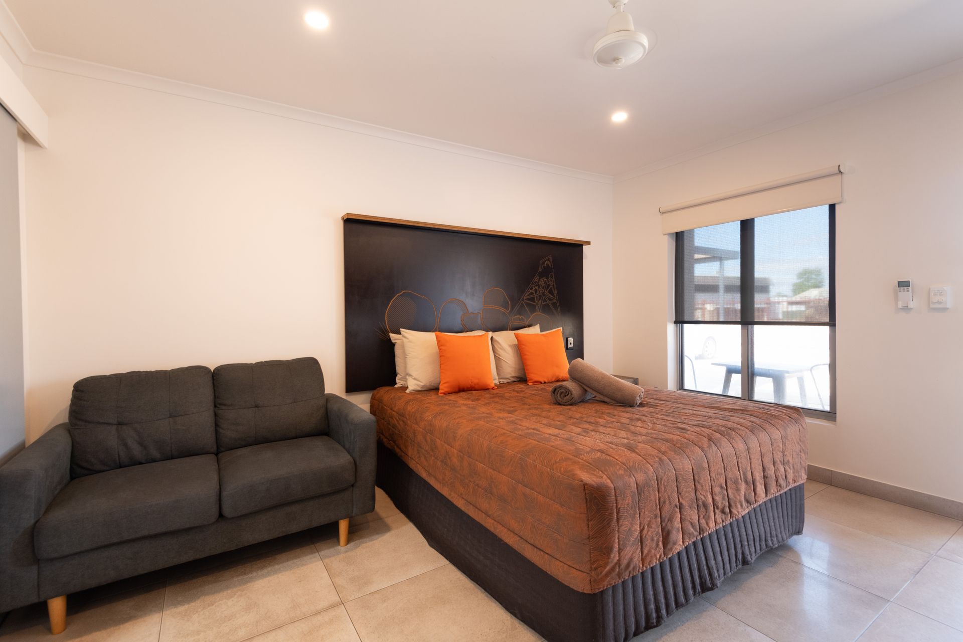 Bedroom with a bed, a small sofa, and a window. Brown and orange bedding, neutral walls.