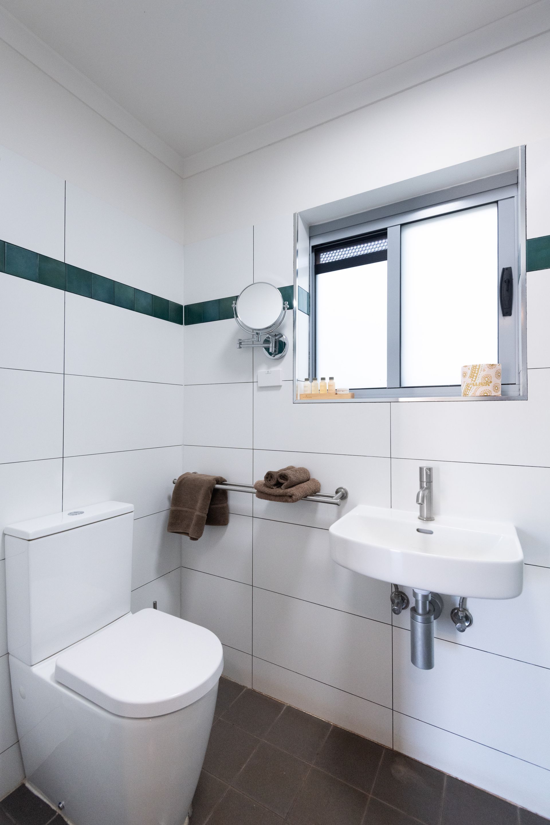 Small, modern bathroom with white tiled walls, toilet, sink, and a small window. Green stripe detail on walls.