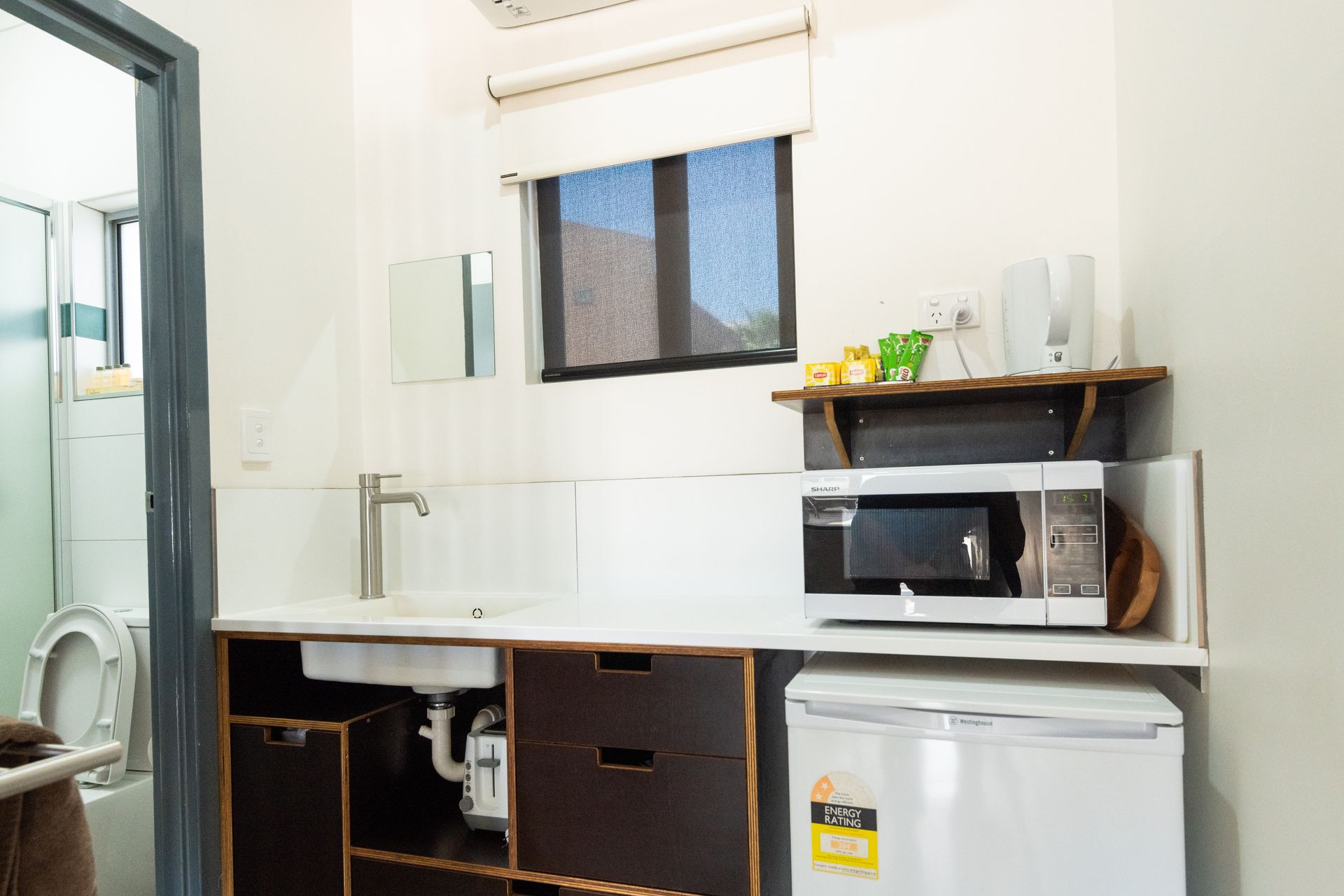 Small kitchen area with a microwave, refrigerator, sink, and a small window. A bathroom is visible on the left.