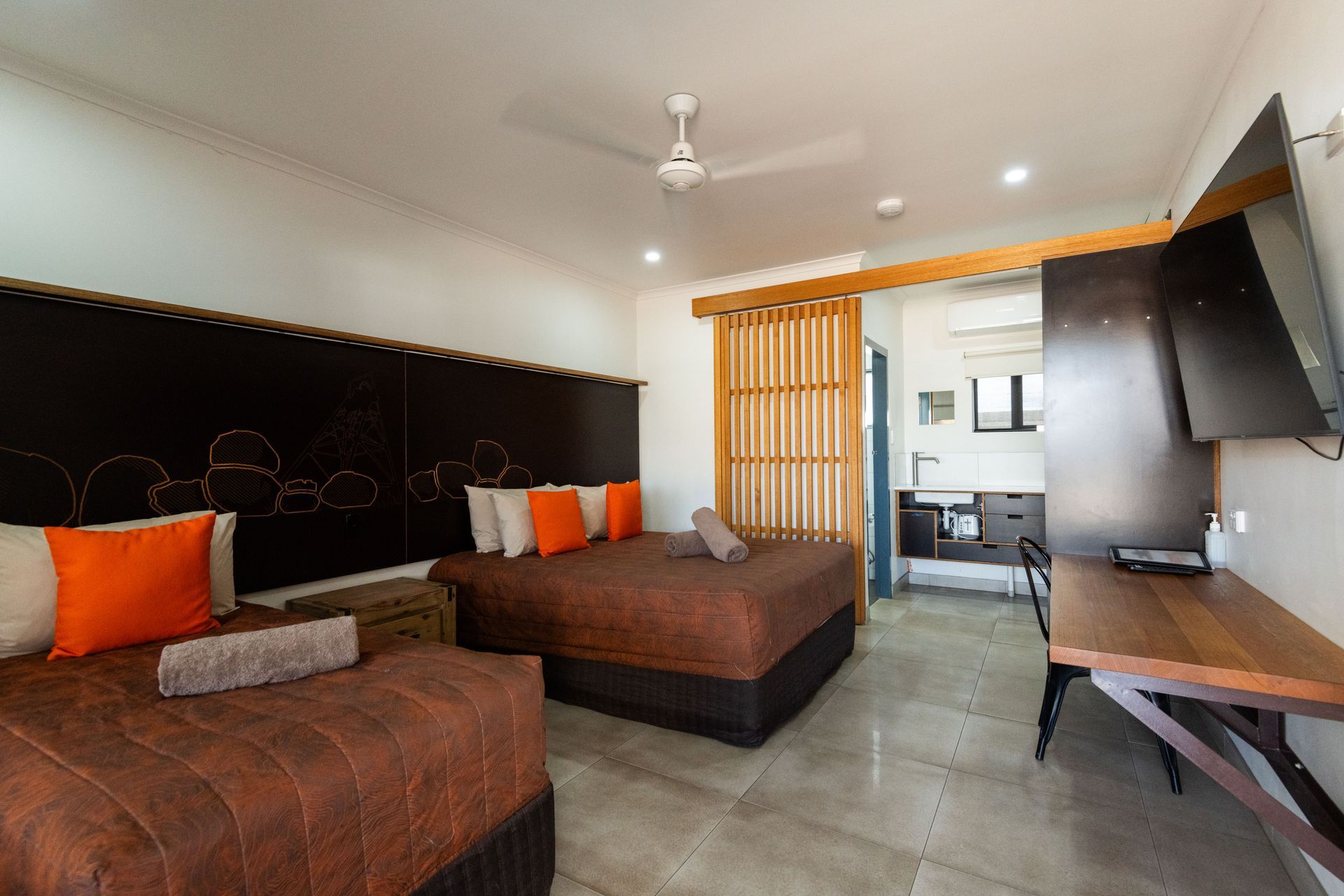 A hotel room with two beds, an orange pillow, a TV, and a small kitchenette area separated by a wooden screen.