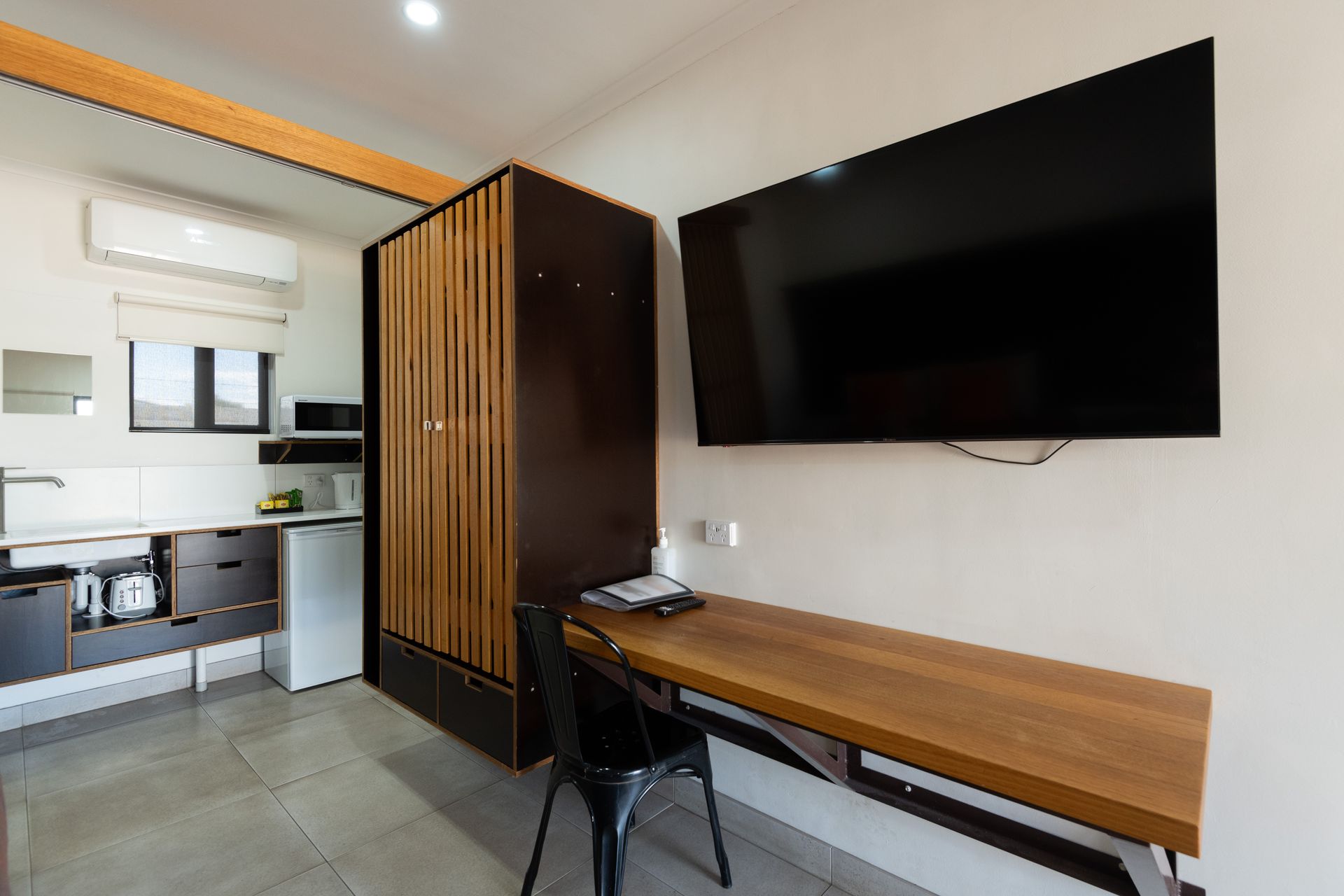 A small studio apartment with a kitchenette, wardrobe, desk, and a large TV mounted on the wall.