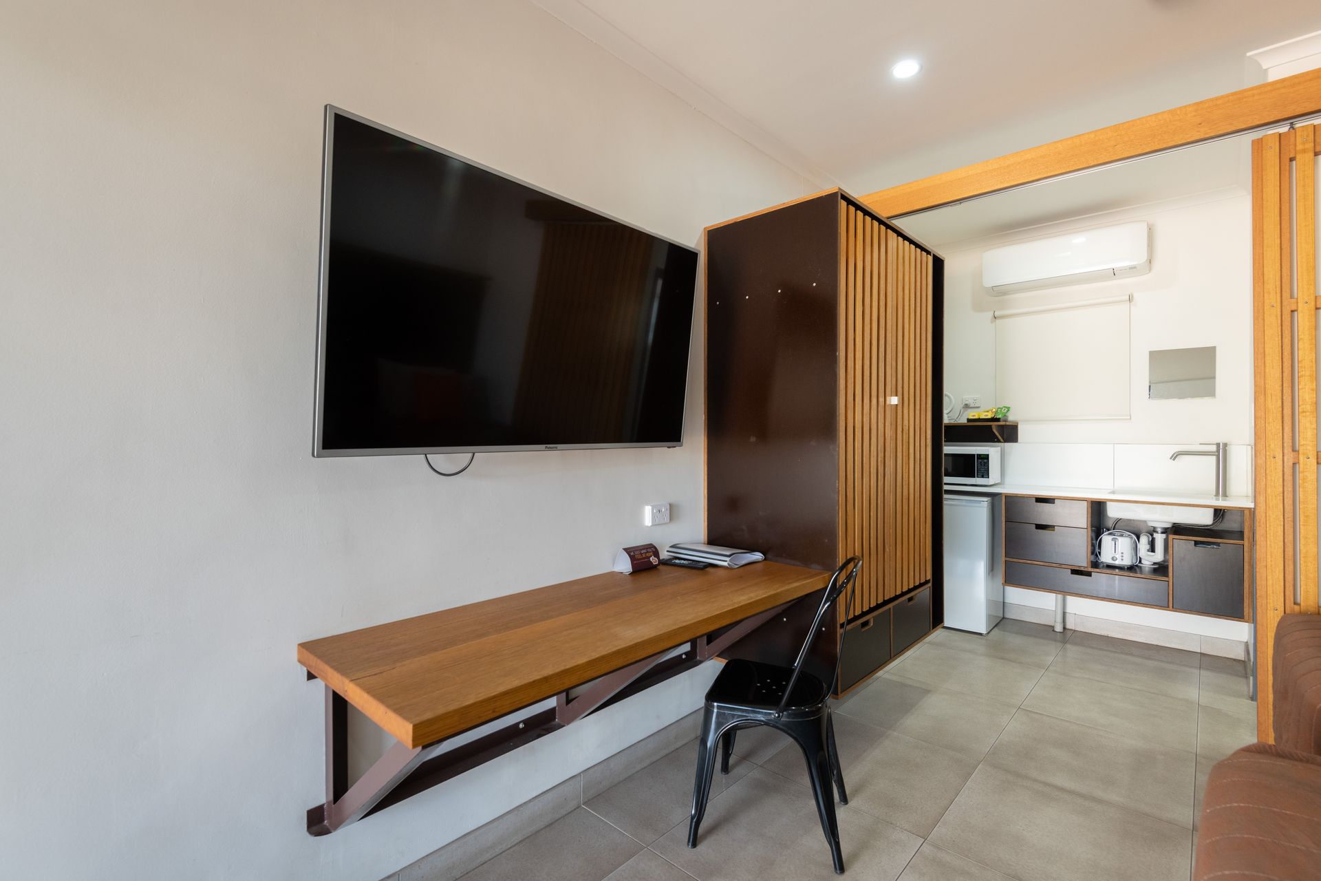 Small studio apartment with mounted TV, fold-down desk, and kitchenette. Brown wood tones and neutral walls.