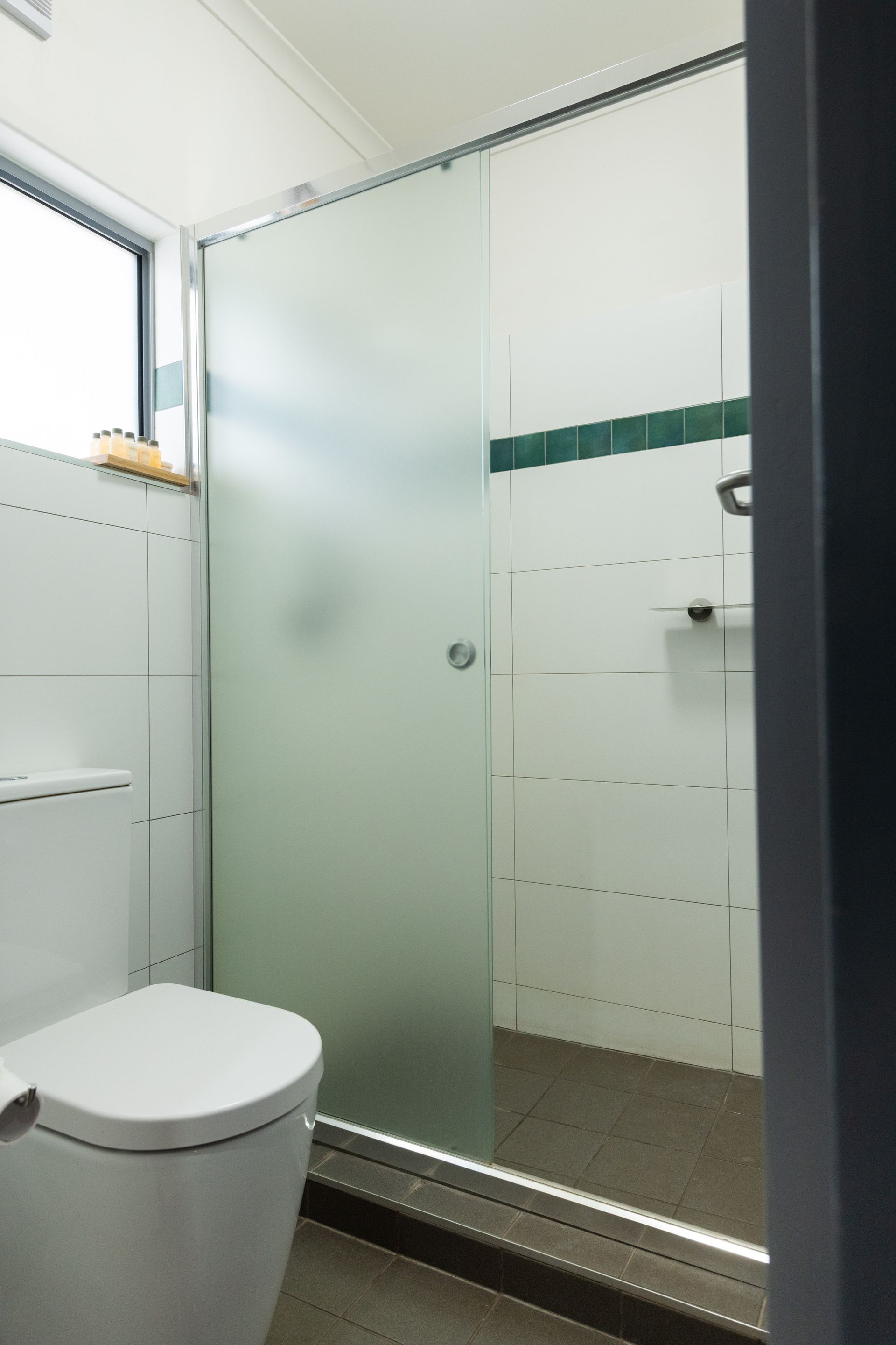 A bathroom with a frosted glass shower, toilet, and window. The walls are white with a green stripe.