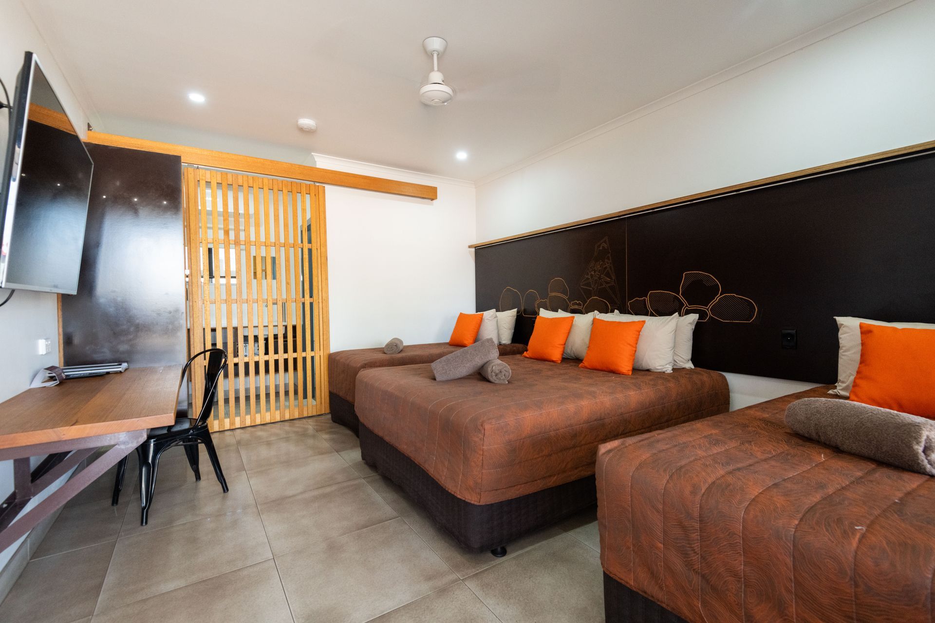 A hotel room with three beds, orange pillows, and a desk. A wooden divider separates a space.