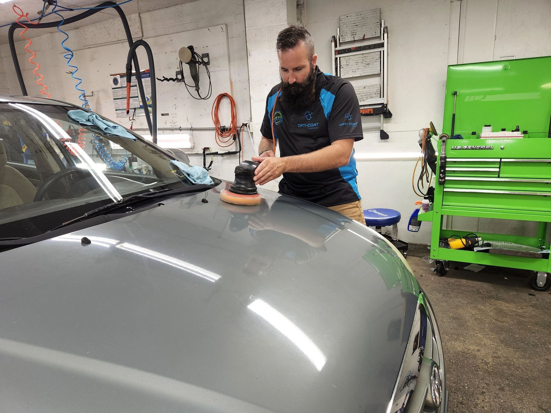 Man polishing a gray car's hood with a machine. He's in a garage, near a tool chest.
