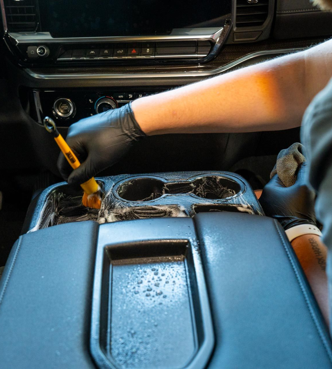Person cleaning a car's center console with a brush. Black gloves, interior shot.