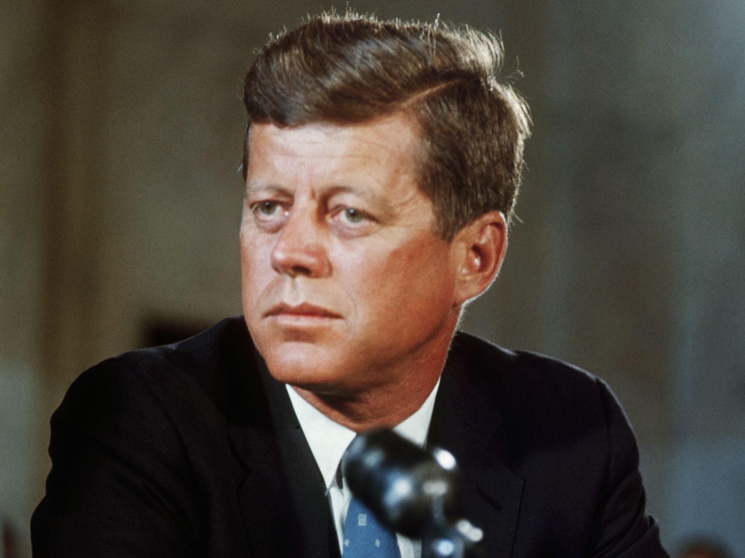 John F. Kennedy at a podium, wearing a dark suit and tie, looking serious with a microphone.