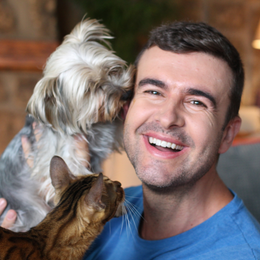 Man smiling while a small dog licks his face and a cat looks up at him indoors