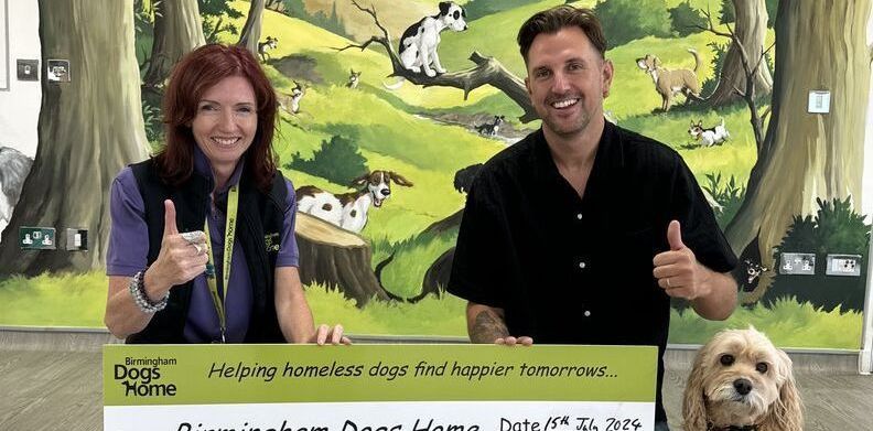 Two people and a dog give a thumbs-up in front of a Birmingham Dogs Home mural, holding a donation sign.