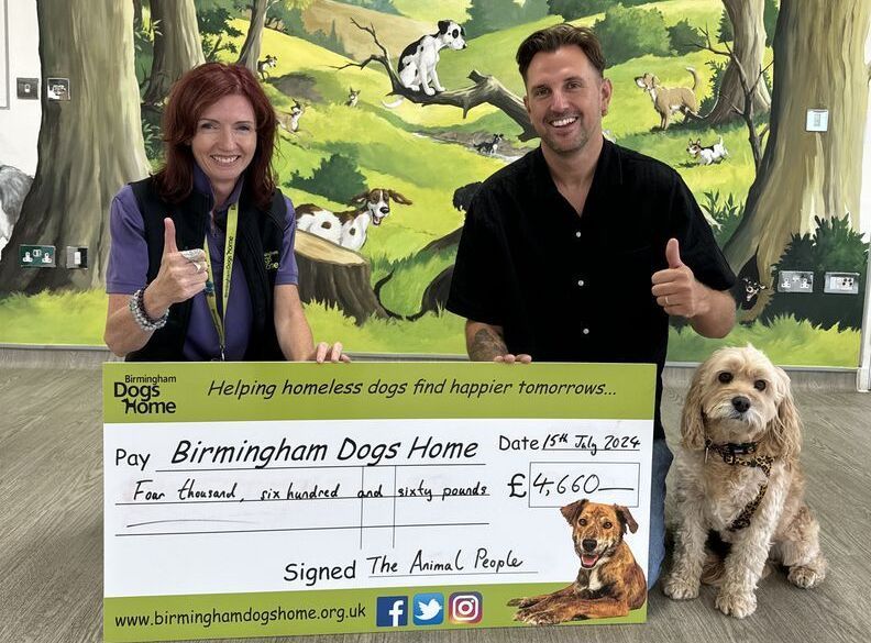 Two people pose behind a giant charity check for Birmingham Dogs Home, with a dog beside them and a dog mural behind.