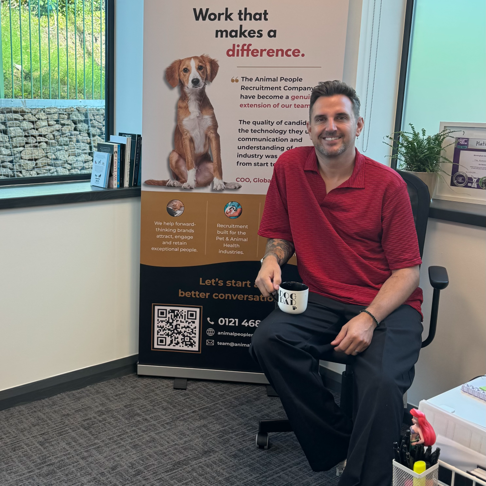 Man in red shirt seated indoors beside a dog-themed poster and QR code display.