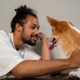 Person lying face-to-face with a small tan-and-white dog, both looking at each other indoors