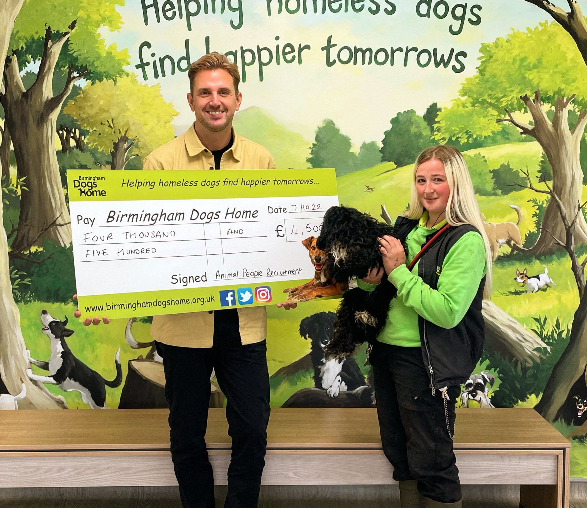 Two people pose with a giant check and a black dog in front of a wall reading “Helping homeless dogs find happier tomorrows.”