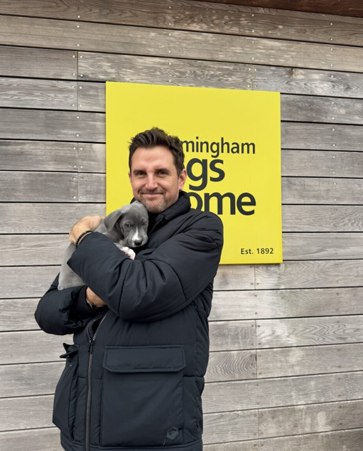 A smiling person in a dark coat holds a small gray puppy in front of a yellow Birmingham Dogs Home sign.