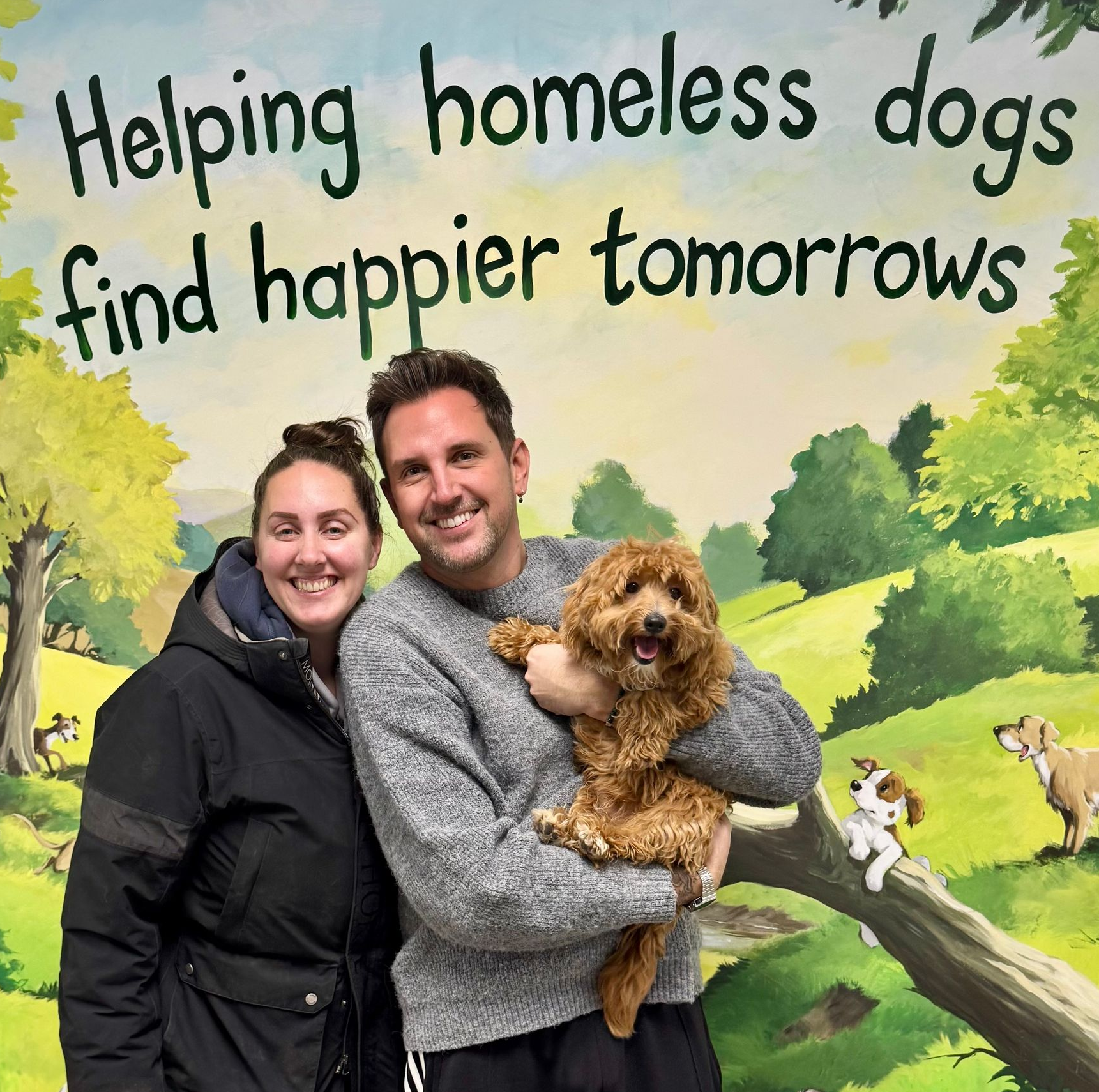 A woman and a man stand smiling in front of a painted mural that says 