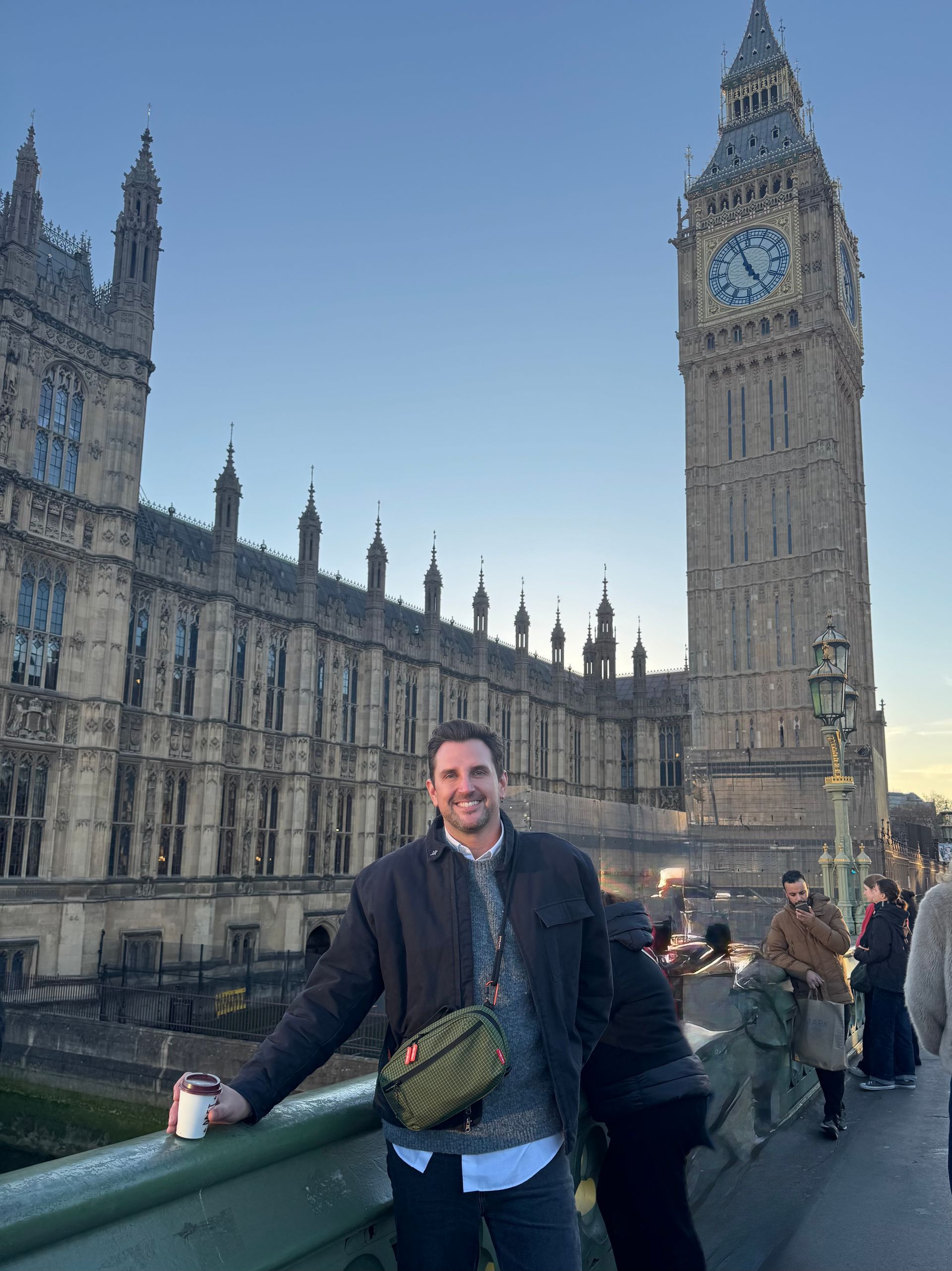 A man in a jacket stands on a bridge with Big Ben and the Houses of Parliament in the background on a clear day.