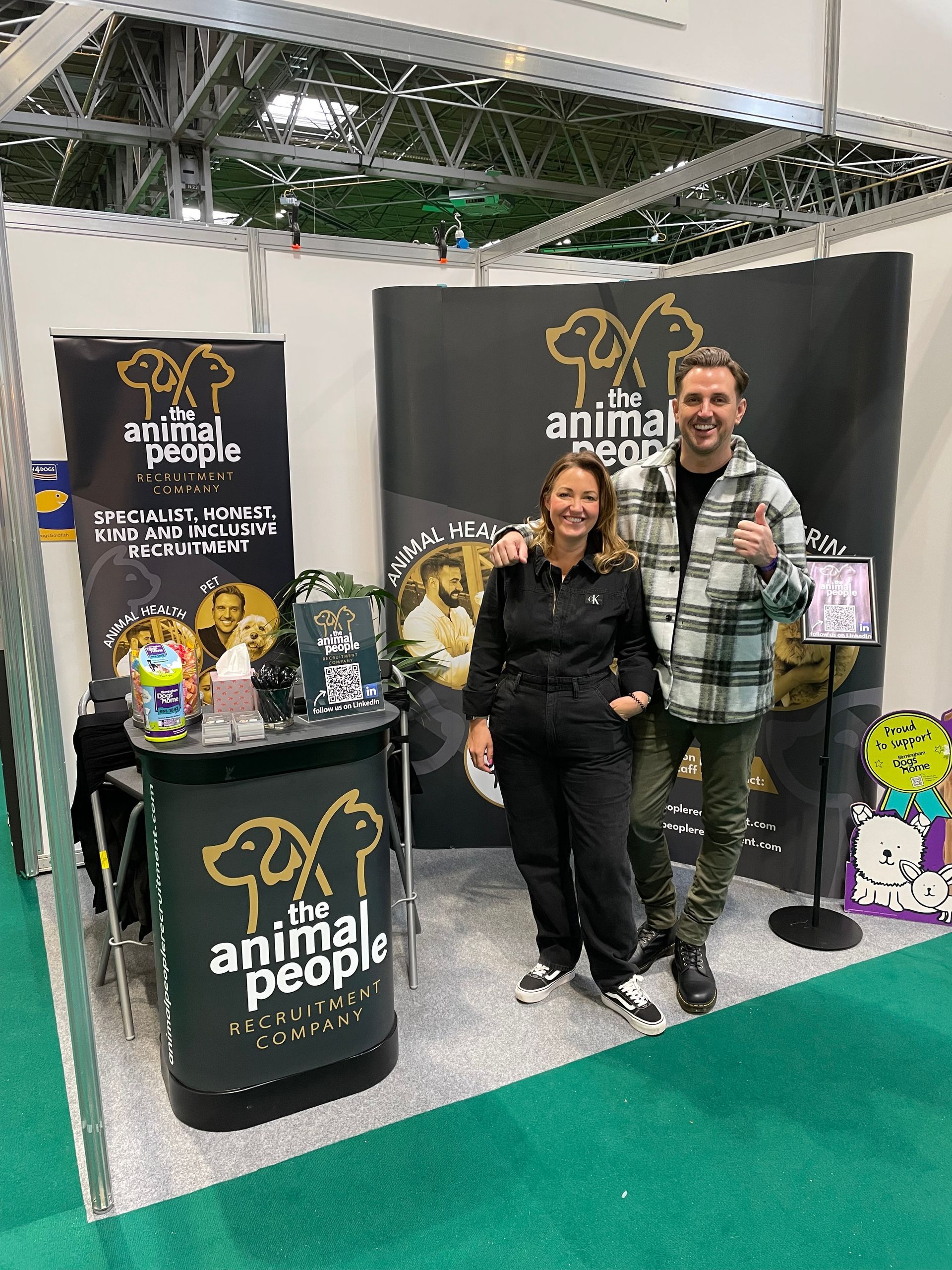 Two people standing at an Animal People booth with black banners and plush dogs at a trade show.