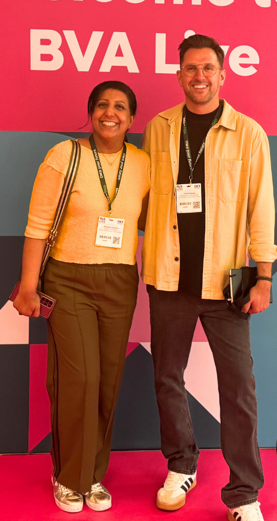 Two smiling people pose in front of a pink BVA Live backdrop, wearing event lanyards.