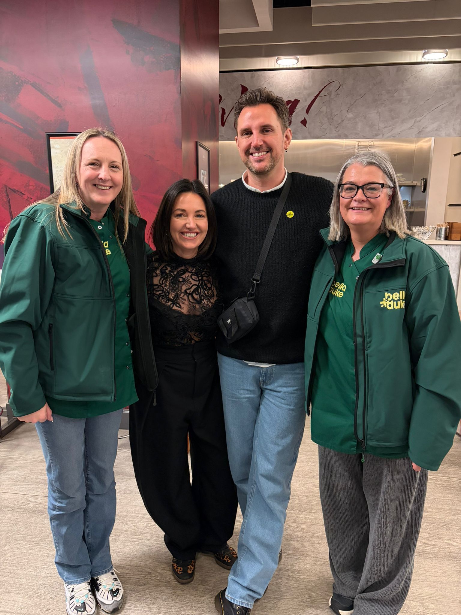 Four people stand side-by-side indoors, smiling toward the camera. They wear casual clothing including green jackets.