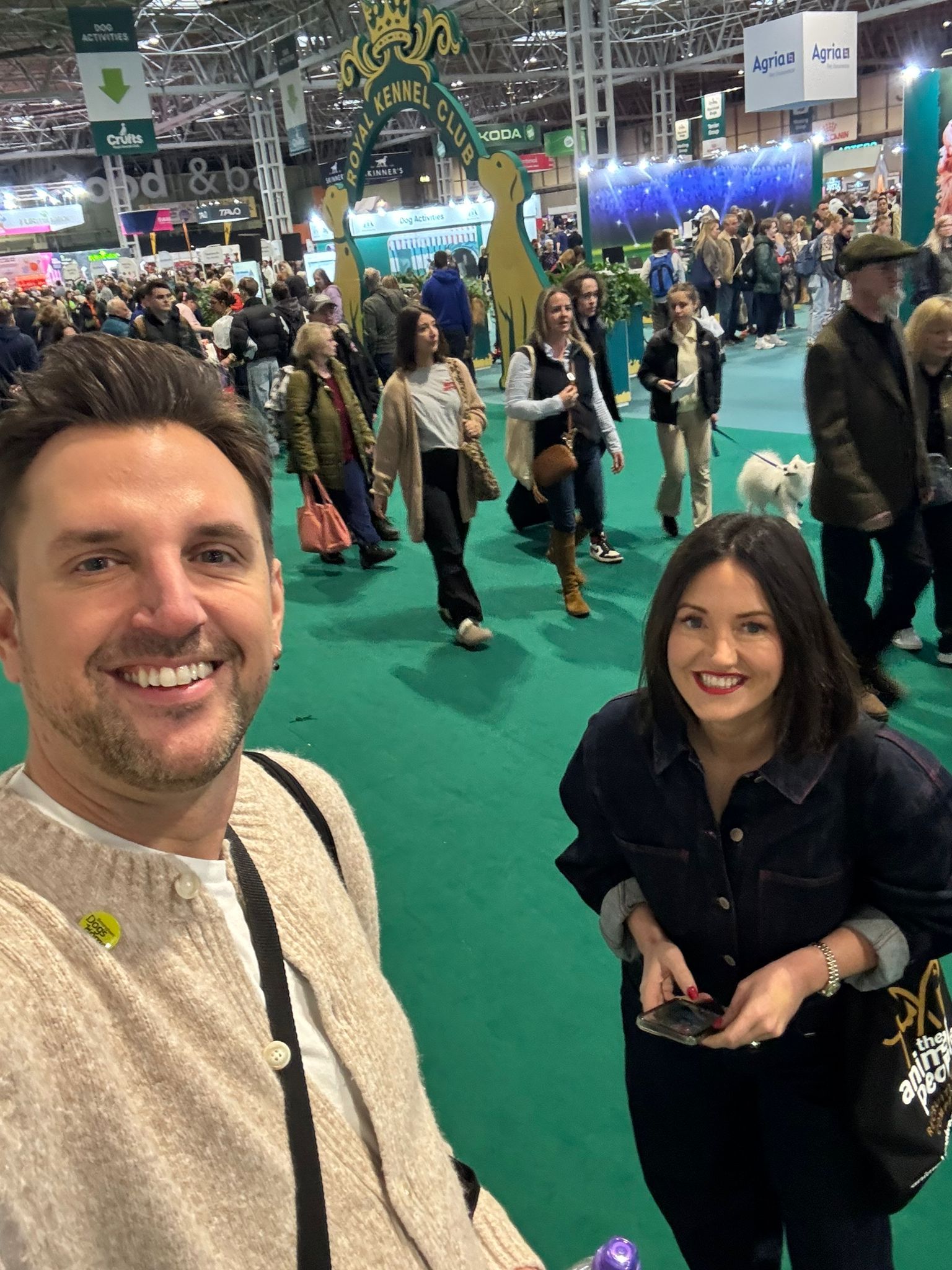 A smiling man and woman pose for a selfie at a crowded indoor event with green carpeting and decorated stalls.
